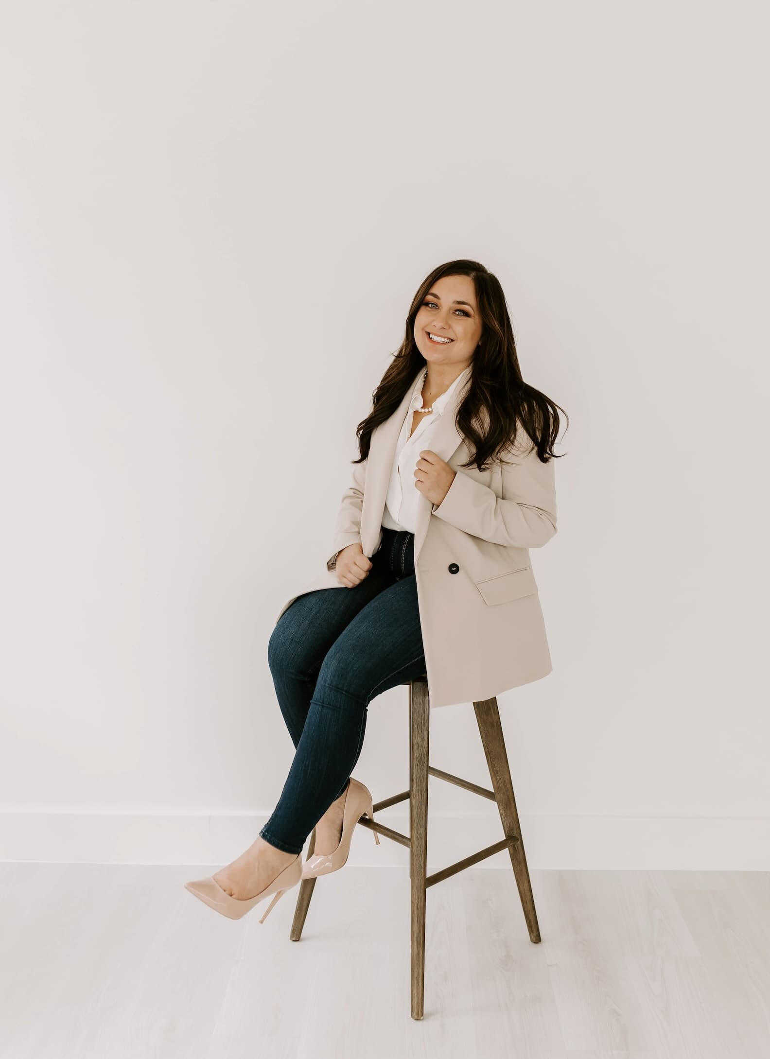 Allie headshot professional brunette woman smiling sitting on stool wearing tan blazer and dark denim