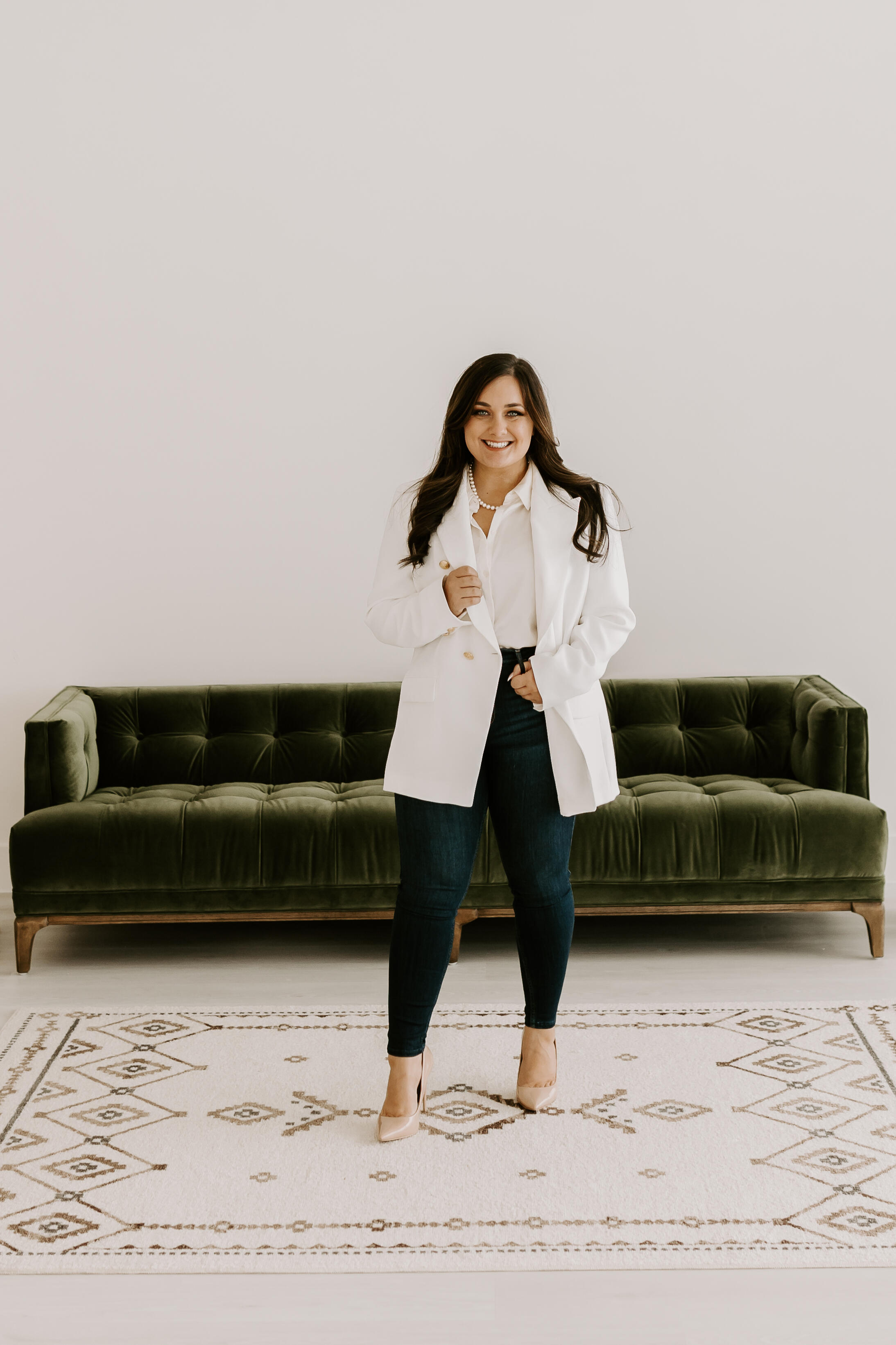 Allie headshot professional brunette woman smiling standing in front of green velvet couch wearing white blazer and dark denim