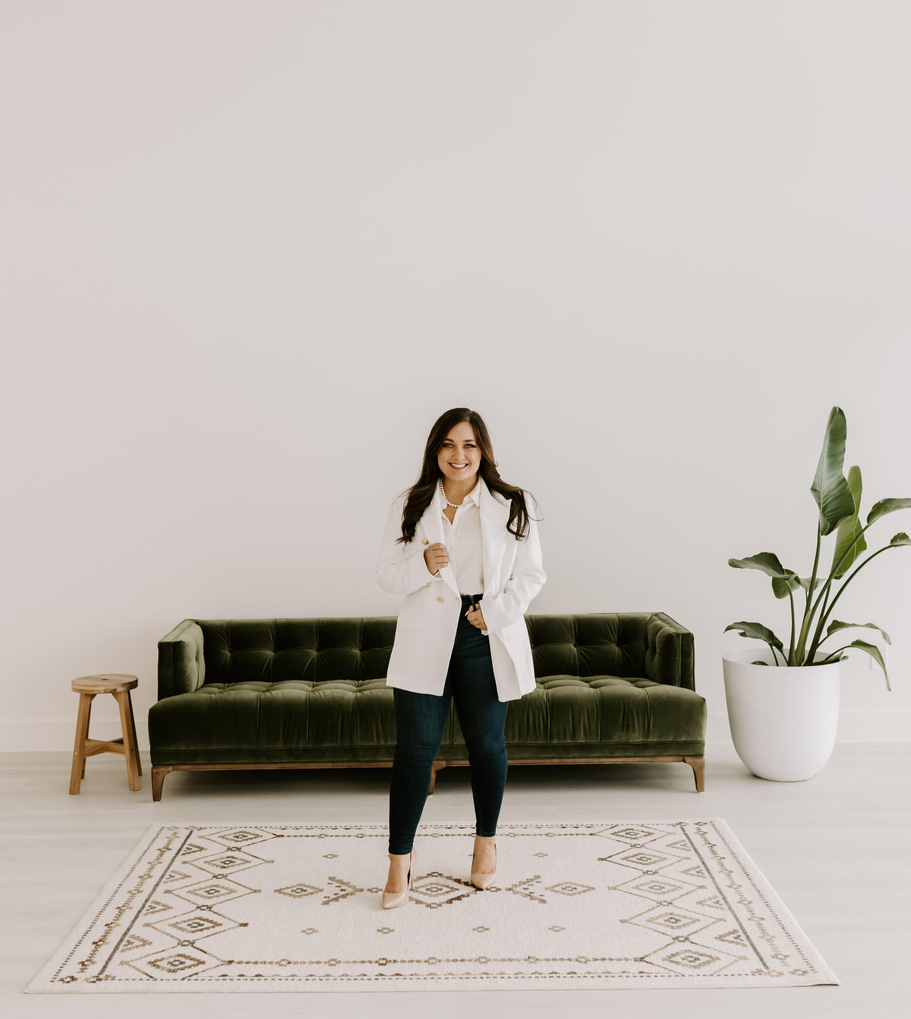 confident smiling woman wearing white blazer and dark denim standing in front of green couch and on boho rug